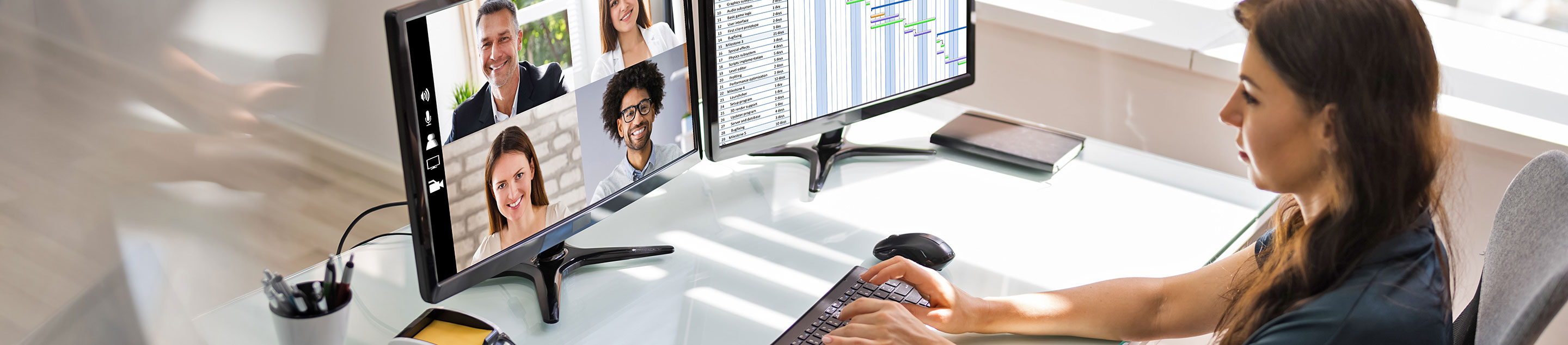 A woman typing at a desk, while attending a video conference meeting with four other participants on her computer screen.